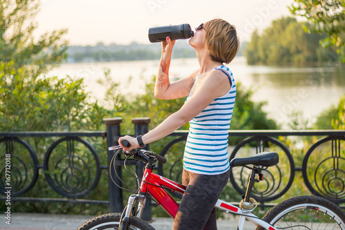 Woman rides a bicycle and drinks from a sports bottle on an alley near the river