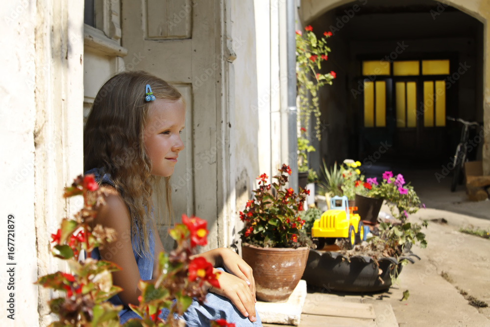 beautiful blonde little girl wearing jeans dress, sitting on a porch in an old garden
