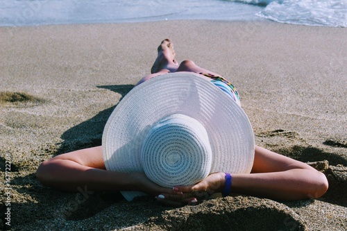 Fototapeta Naklejka Na Ścianę i Meble -  The woman lies on the sand against the background of the sea surf in a white hat with wide brim.