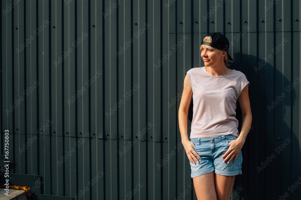 Young stylish blonde woman in shorts and black cap in front of black wall. Urban concept. Pretty hip hop girl outdoors Young stylish blonde woman in shorts and black cap in front of black wall. Urban concept. Pretty hip hop girl outdoors