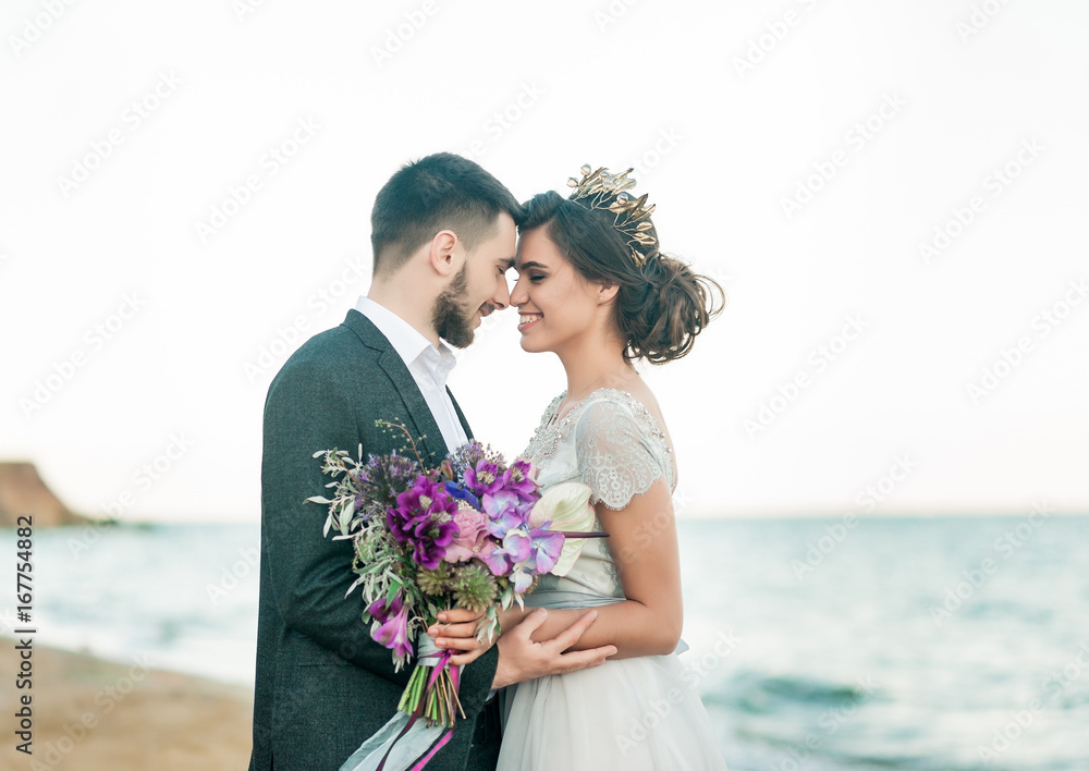 Wedding bride and groom with bouquet posing near sea on sunset