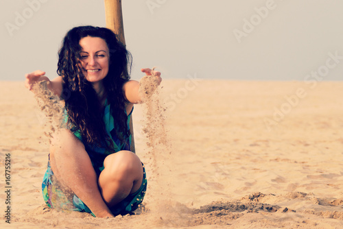 Happy young girl in a blue dress is sitting on the sand on the beach. Toned.