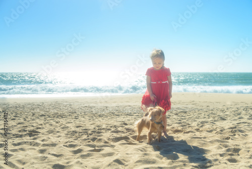 Little smiling blond girl in red dress with dog on the beach