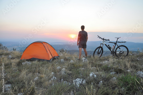 The traveler has set up a tent and enjoys the sunset. A cyclist with a ranch next to the tent is preparing for the night