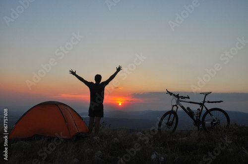 The traveler has set up a tent and enjoys the sunset. A cyclist with a ranch next to the tent is preparing for the night