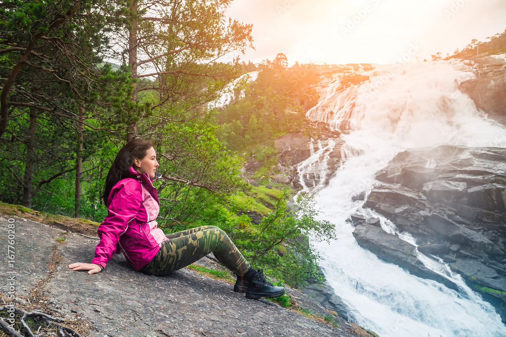 Naklejka premium A young hiker girl sitting on the stone and admires a waterfall in the Valley of the waterfalls Norway