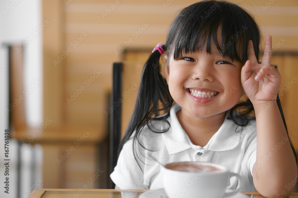 Asian children cute or kid girl white teeth smile and hot cocoa or chocolate drinking for breakfast and show two fingers because delicious in the morning on wood table in the home or cafe restaurant Asian children cute or kid girl white teeth smile and hot cocoa or chocolate drinking for breakfast and show two fingers because delicious in the morning on wood table in the home or cafe restaurant