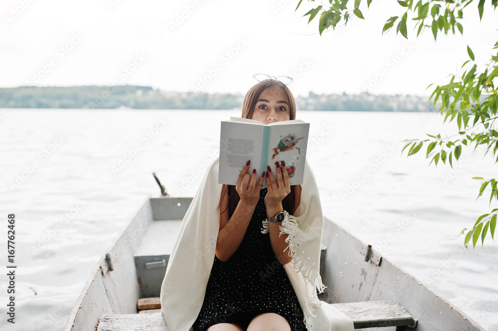 Obraz premium Portrait of an attractive woman wearing black polka dots dress, white shawl and glasses reading a book in a boat on a lake.
