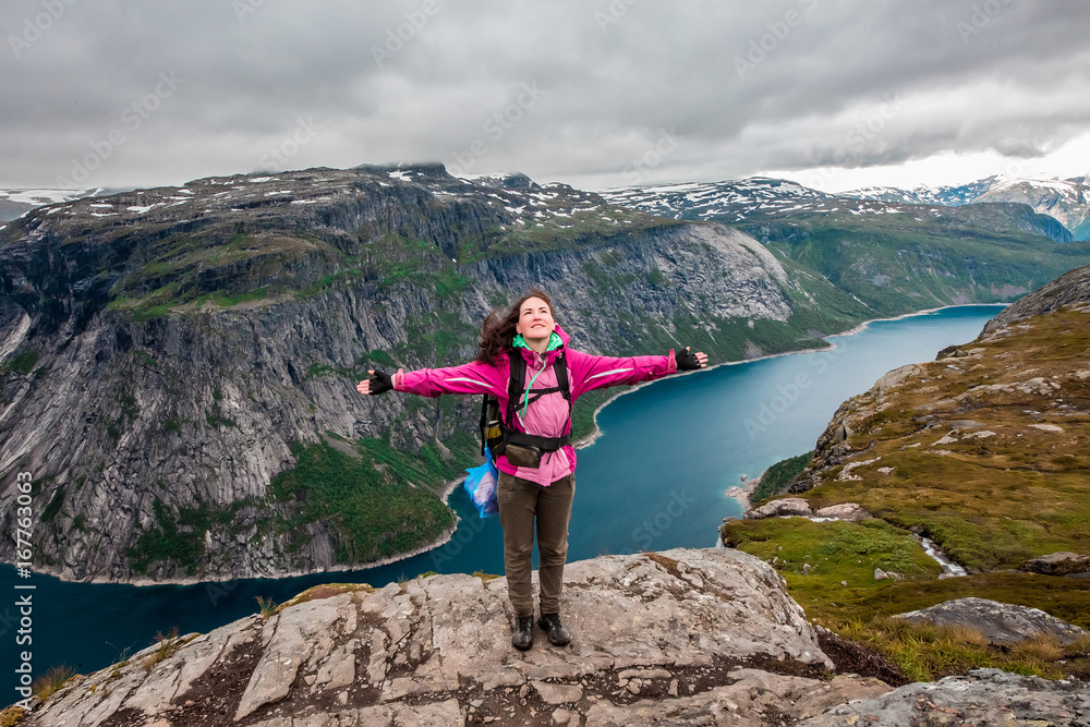 Obraz premium Young hiker girl with backpack enjoying valley view from top of a mountain with open arms. High resolution panorama, Norway. Troltunga route