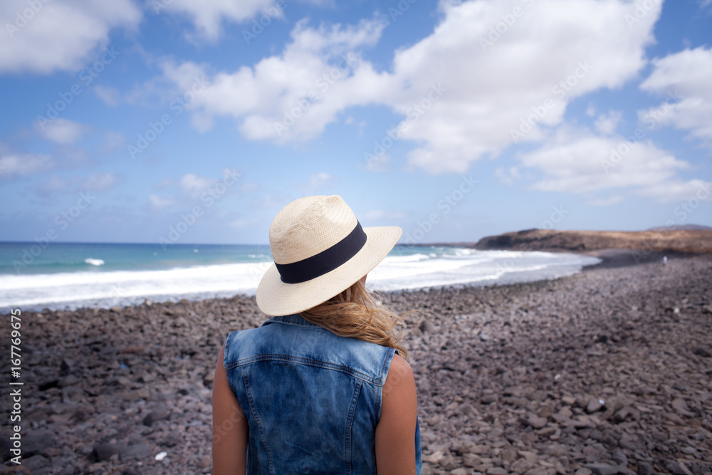 woman back with hat. looking over the sea. standing on a stones beach. Lanzarote Canary Island 