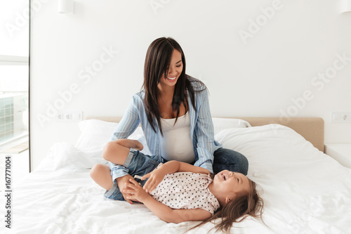 Smiling asian mother playing with her little daughter on bed
