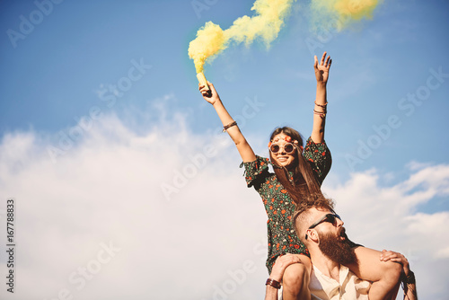 Young boho woman holding yellow smoke flare on boyfriend's shoulders at festival photo