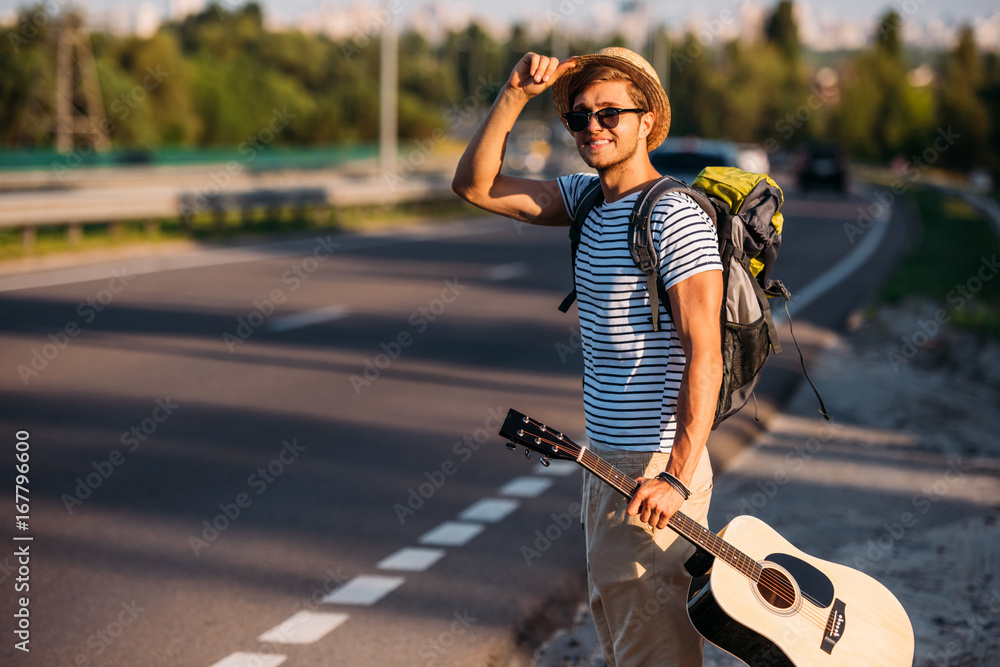 smiling man with backpack and guitar in hand hitchhiking alone down road