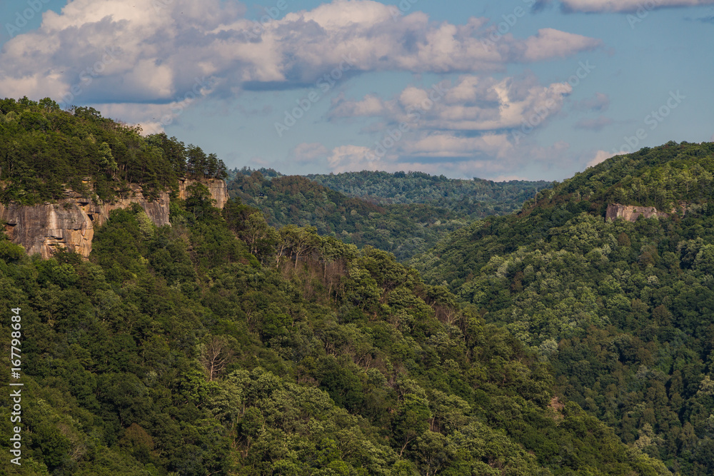 Southern West Virginia mountain tops with rock outcrops visible through the green trees of summertime and a cloudy blue sky overhead.