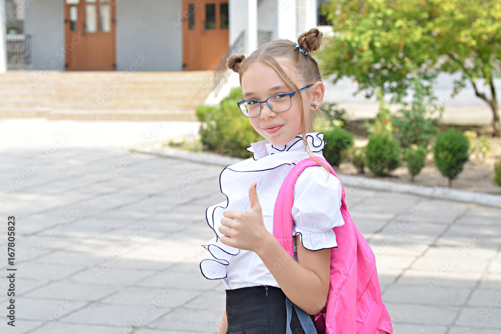 Fototapeta premium Smiling cute schoolgirl of primary school in glasses. Girl with a pink bag near the building of the school outdoors.