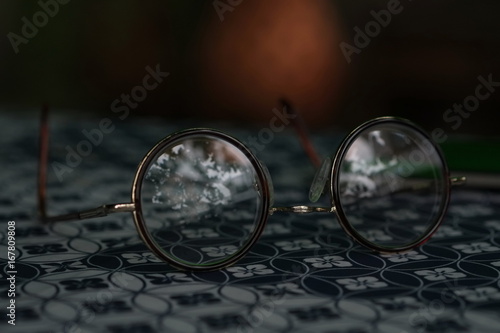 Eyeglass with a circular frame placed on black patterned table with a reflection reflect the surrounding on the lens of the eyeglass.