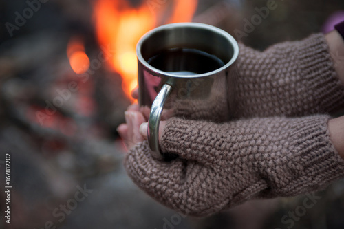 Hands in mitts holding hot tea cup outdoor near bonfire
