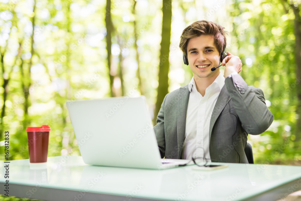 Smile handsome Businessman with headset sitting at the office desk with laptop computer and cup of coffee in call center in green forest park. Business concept. Smile handsome Businessman with headset sitting at the office desk with laptop computer and cup of coffee in call center in green forest park. Business concept.