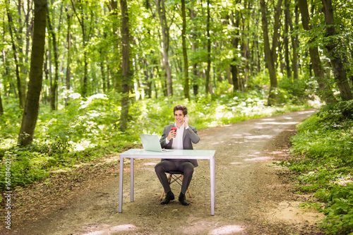 Businessman sitting at the office desk with laptop computer and cup of coffee talking mobile phone in green forest park. Business concept.