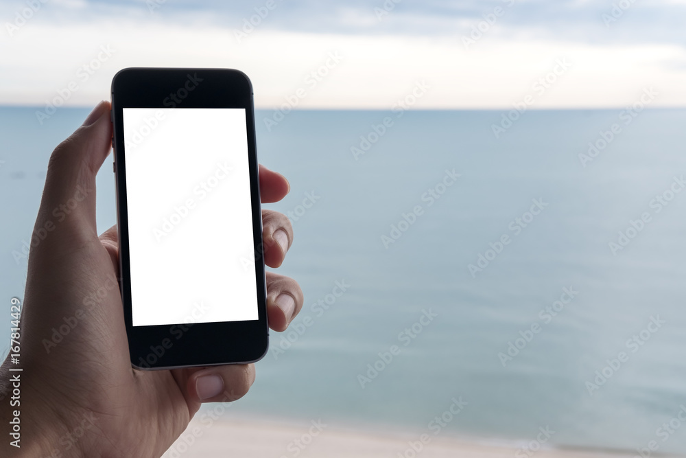 Mockup image of a hand holding black mobile phone with blank white screen with blue sky , white beach and the sea in background
