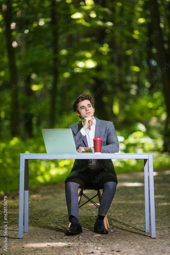 Thinking Businessman with hands on chin sitting at the office desk work at laptop computer and cup of coffee on road of green forest park. Business concept. Thinking Businessman with hands on chin sitting at the office desk work at laptop computer and cup of coffee on road of green forest park. Business concept.