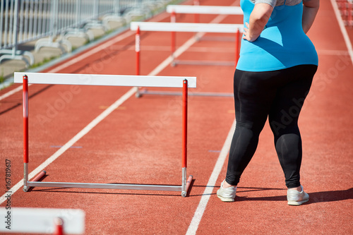 Determined plus-size female in activewear standing by hurdle on racetrack