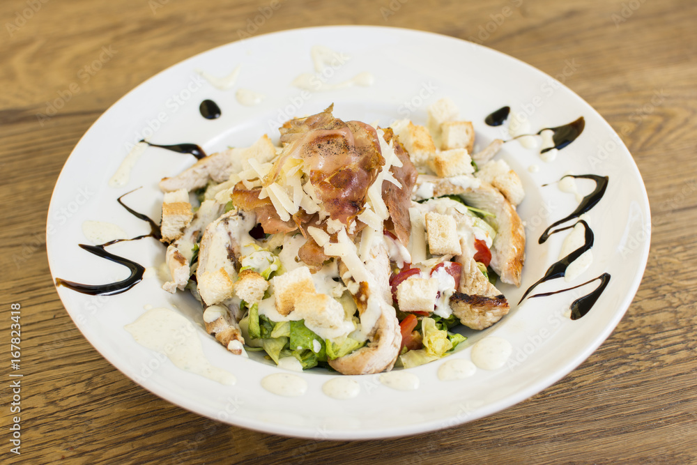 Healthy grilled chicken Caesar salad with cheese and croutons. Bacon and lettuce slices in a white plate on a wooden table. Healthy grilled chicken Caesar salad with cheese and croutons. Bacon and lettuce slices in a white plate on a wooden table.