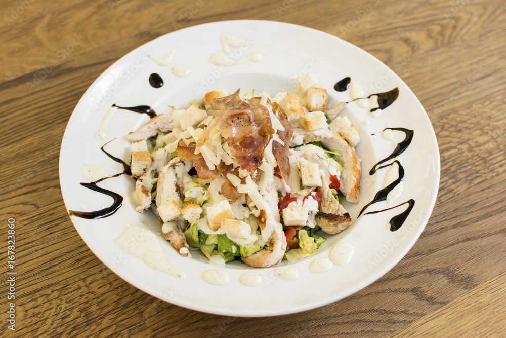 Healthy grilled chicken Caesar salad with cheese and croutons. Bacon and lettuce slices in a white plate on a wooden table. Healthy grilled chicken Caesar salad with cheese and croutons. Bacon and lettuce slices in a white plate on a wooden table.