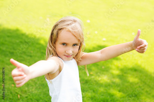 Happy little girl showing thumbs up  outdoors in summer day.