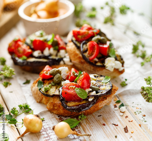 Bruschetta with grilled aubergine, cherry tomatoes, feta cheese, capers and fresh aromatic herbs, on a wooden table. Delicious Mediterranean appetizer photo