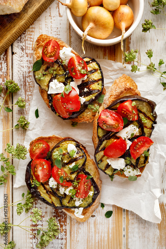Bruschetta with grilled aubergine, cherry tomatoes, feta cheese, capers and fresh aromatic herbs on a wooden table, top view. Delicious Mediterranean appetizer