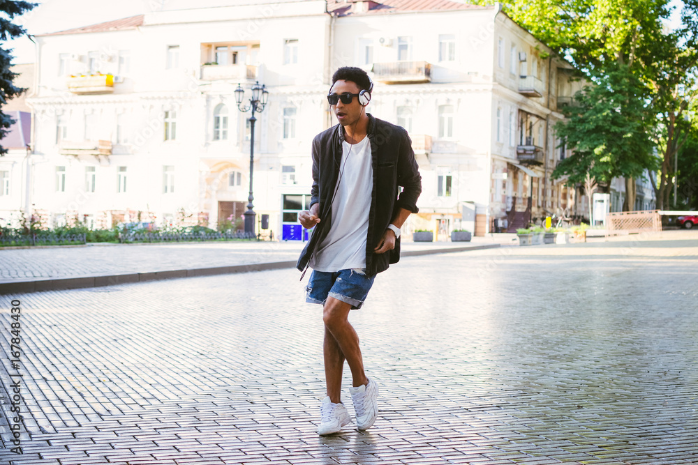 Portrait of young stylish hipster black man in white headphones and sun glasses dancing outdoor in city centre, having some fun