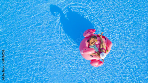 AERIAL TOP DOWN: Young man and woman drinking cocktails and relaxing while lying on fun inflatable pink flamingo floatie. Sexy couple in swimsuit sipping drinks, enjoying floating on sunny pool water