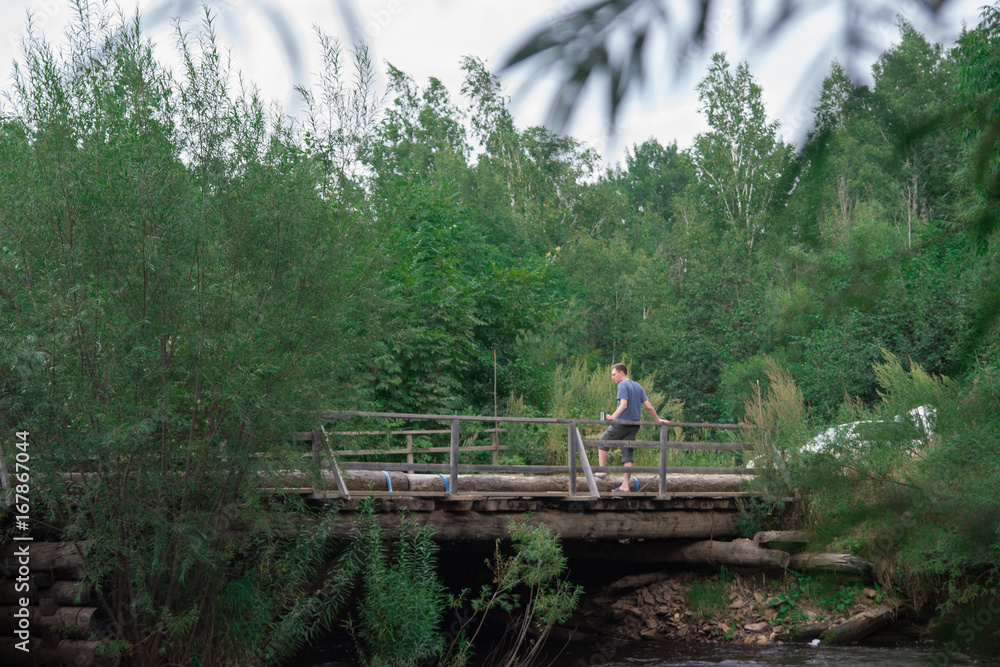 Man sitting on the wooden bridge railings on bridge over the small village river Man sitting on the wooden bridge railings on bridge over the small village river