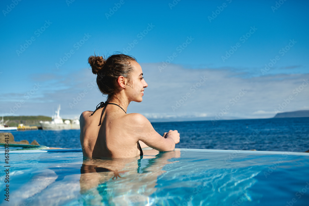 Young cheerful girl swimming in water of pool looking at the water on background of sea, Iceland, West Fjords. back view