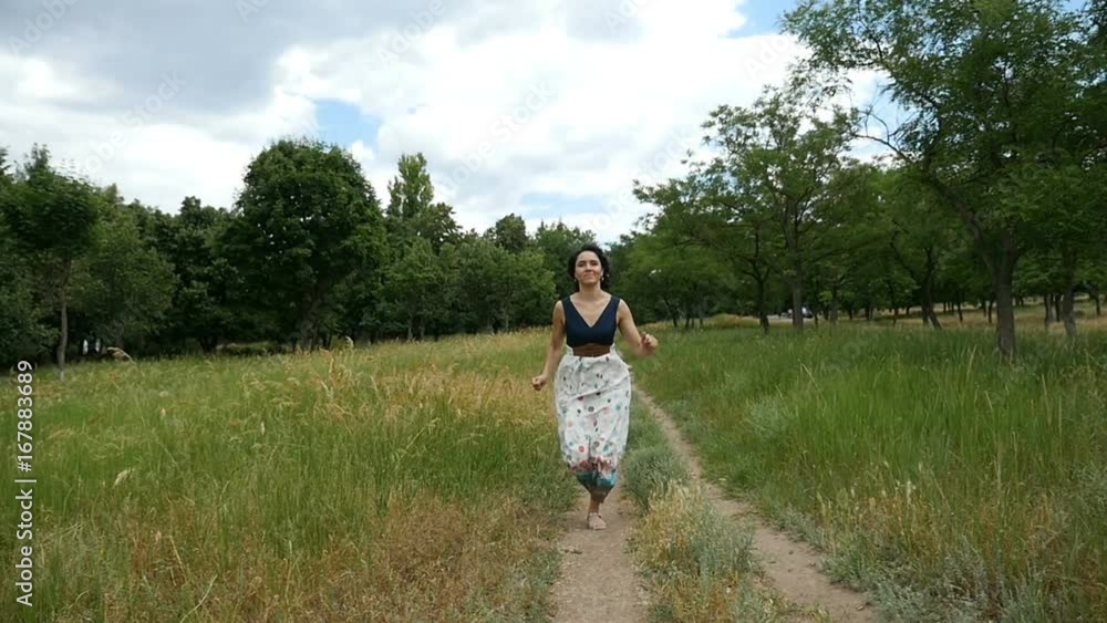 A charming brunette woman, in a white dress with black top and open shoulders runs along a country road and smiles happily in slow motion. She is on picturesque lawn with green trees on a sunny day A charming brunette woman, in a white dress with black top and open shoulders runs along a country road and smiles happily in slow motion. She is on picturesque lawn with green trees on a sunny day