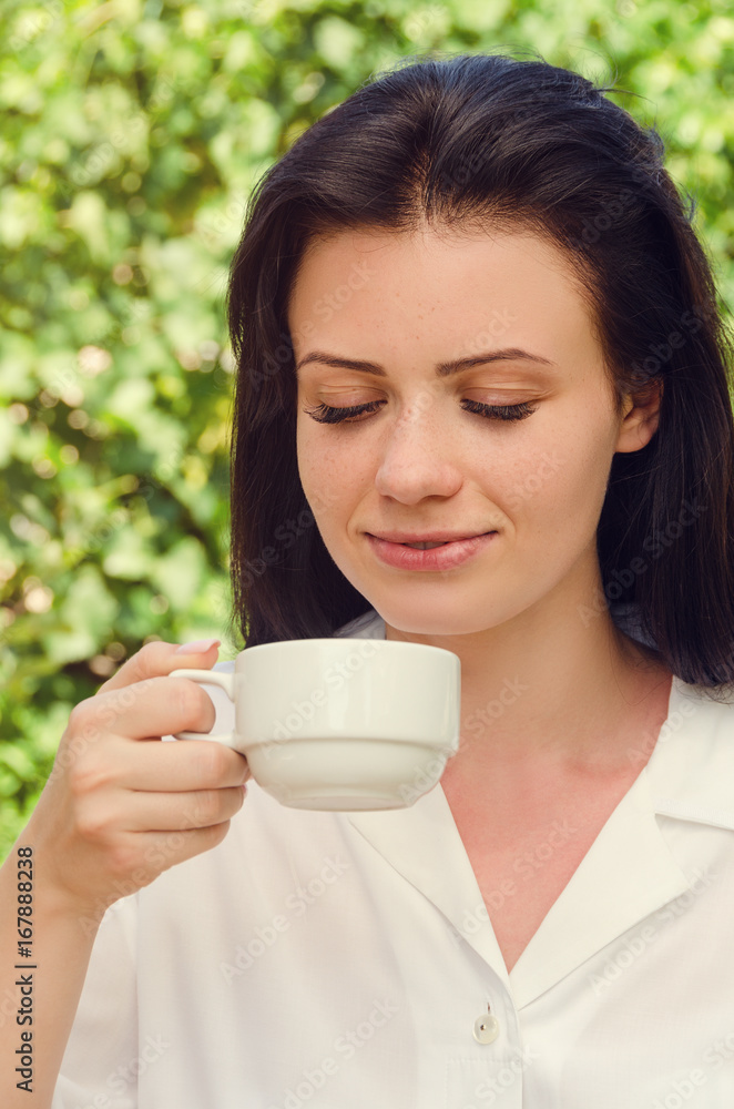 Young beautiful woman in white shirt smiling and drink coffee. Portrait. Summer green park. Young beautiful woman in white shirt smiling and drink coffee. Portrait. Summer green park.