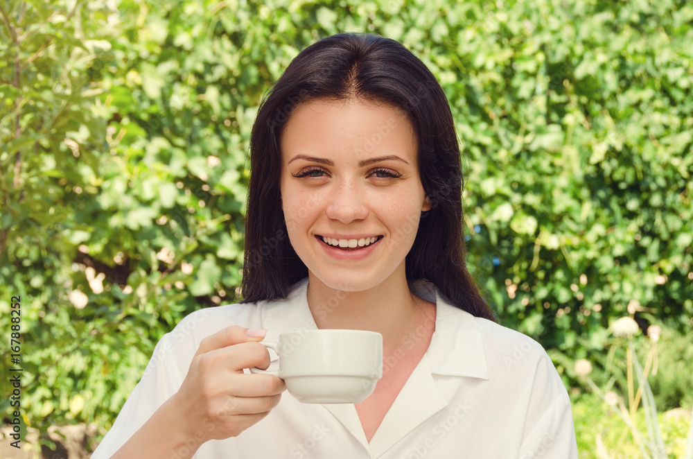Young beautiful woman in white shirt smiling and drink coffee looking at camera. Portrait. Summer green park. Young beautiful woman in white shirt smiling and drink coffee looking at camera. Portrait. Summer green park.