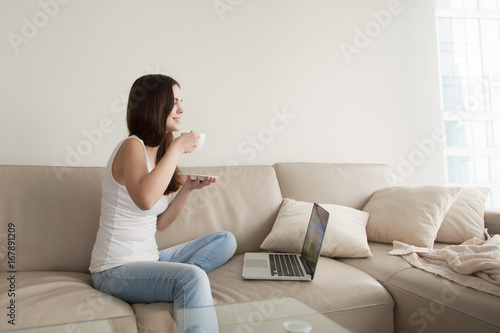 Young woman drinking coffee while sitting comfortably at home sofa, happy teen relaxing with laptop computer, relaxed girl enjoying spending time alone watching series online on pleasant weekend