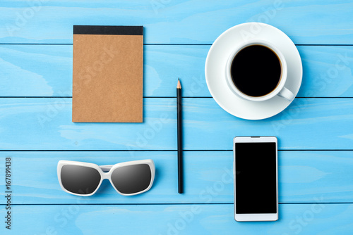Smart phone, sunglasses, notebook and a cup of coffee on blue wooden table