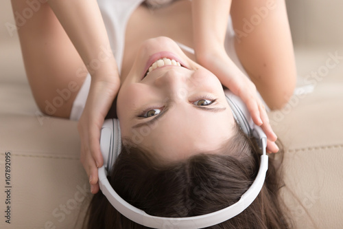 Pretty young woman lying on sofa at home in white headphones, cute teen girl with smiling face listening to audiobook, enjoying favorite mp3 music radio station, close up view, head shot upside down