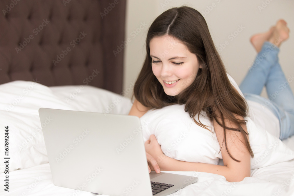 Pretty young lady using laptop while lying on bed in the morning, smiling teen girl checking news feed on pc at home, playing games, browsing web, shopping online, communicating with friends Pretty young lady using laptop while lying on bed in the morning, smiling teen girl checking news feed on pc at home, playing games, browsing web, shopping online, communicating with friends