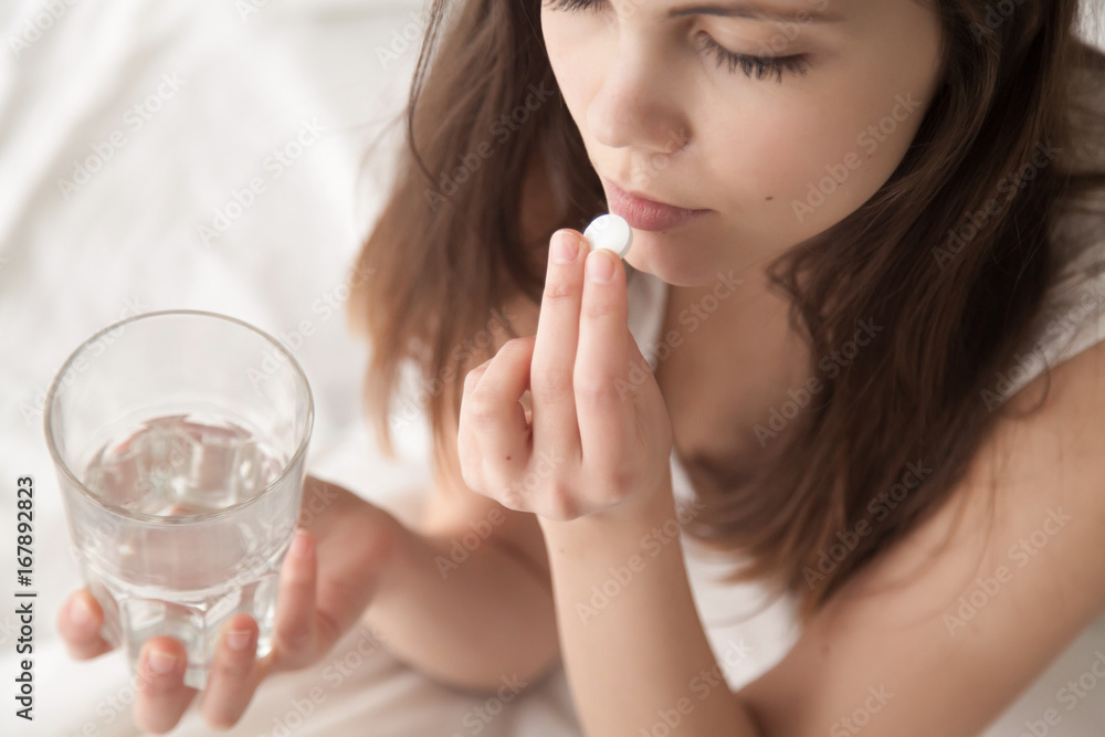Unhealthy sick woman suffers from insomnia or headache, takes sleeping pill while sitting in bed with glass of water, depressed girl holds antidepressant meds, painkiller for menstrual pain, close up Unhealthy sick woman suffers from insomnia or headache, takes sleeping pill while sitting in bed with glass of water, depressed girl holds antidepressant meds, painkiller for menstrual pain, close up
