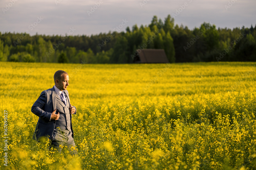 Young and attractive man in a suit standing in yellow rape field and looking somewhere.
