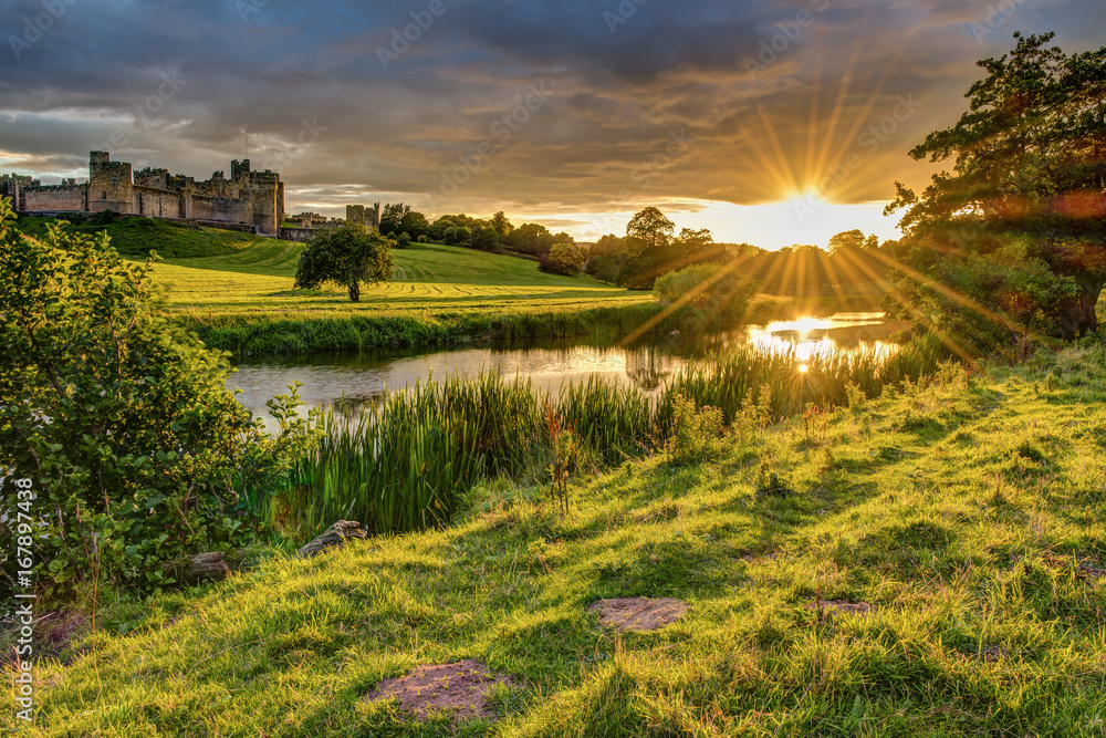 Fototapeta premium Promienie słoneczne nad rzeką Aln w Alnwick / The River Aln biegną przez Northumberland od Alnham do Alnmouth. Widzisz tutaj poniżej Alnwick Town i Castle na panoramie, gdy zachodzi słońce