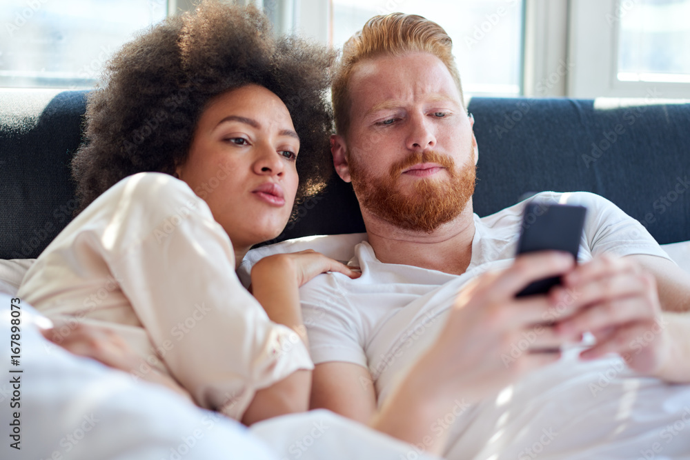 Fototapeta premium Young couple laughing together and using mobile phone while lying in bed