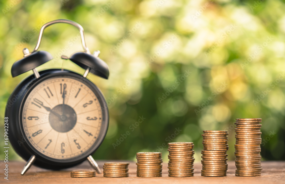 Money coin stack arranged as a graph on wood and black alarm clock with blur nature the scene behind, concept of time to money growth Money coin stack arranged as a graph on wood and black alarm clock with blur nature the scene behind, concept of time to money growth