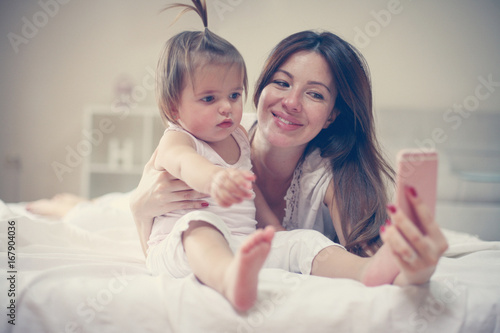 Mother with her little baby having fun in the bed. Mother making self-picture with her cute little baby in the bed.
