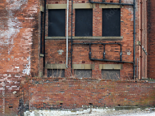 derelict abandoned commercial or factory type building with boarded up windows decaying bricks and a collapsing wall with weeds