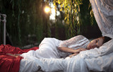 Young pensive woman lies on bed with bedding and baldachin near tree on sunset.